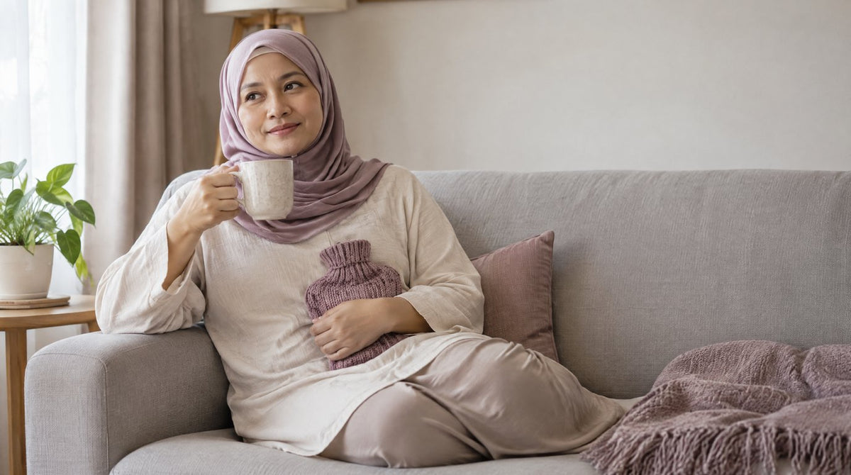 A woman in a hijab resting on a sofa at home with a hot water bottle on her lower tummy and a cup of tea