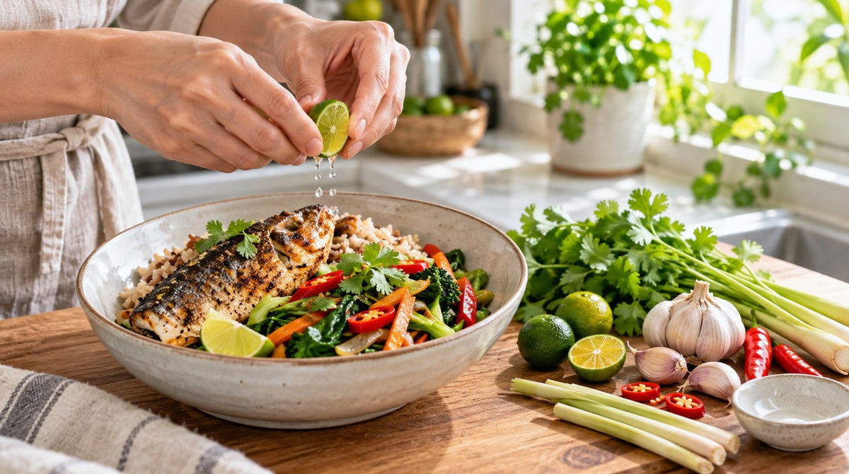 Hands squeezing lime over a bowl of grilled fish, brown rice and vegetables beside garlic, chilli, lemongrass and coriander in a bright kitchen