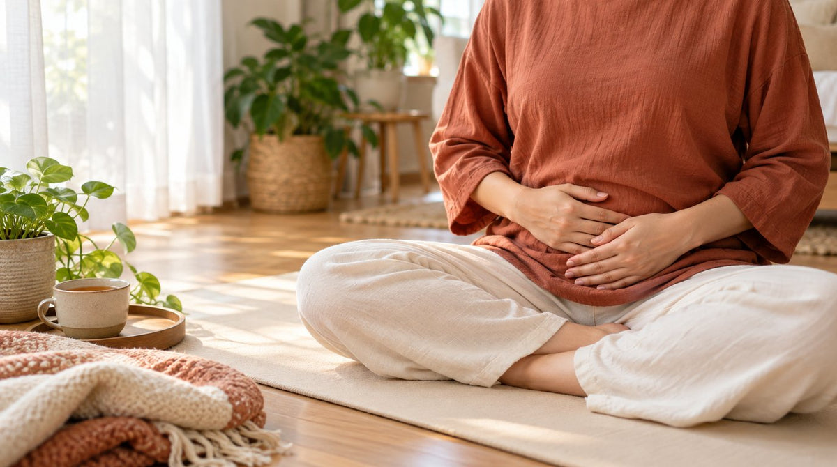 Woman in a terracotta top sitting cross-legged on a yoga mat with hands resting on her lower belly, beside a cup of tea and a blanket
