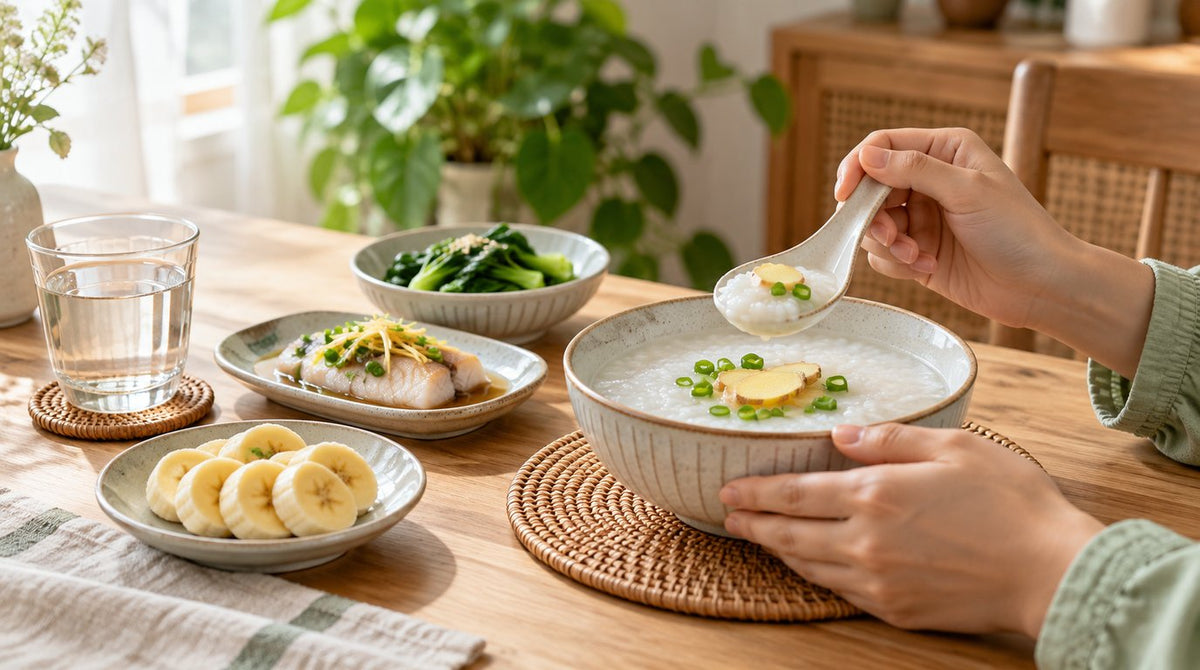 Hands holding a spoon over a bowl of plain rice porridge with ginger, beside steamed fish, green vegetables, banana slices and a glass of water