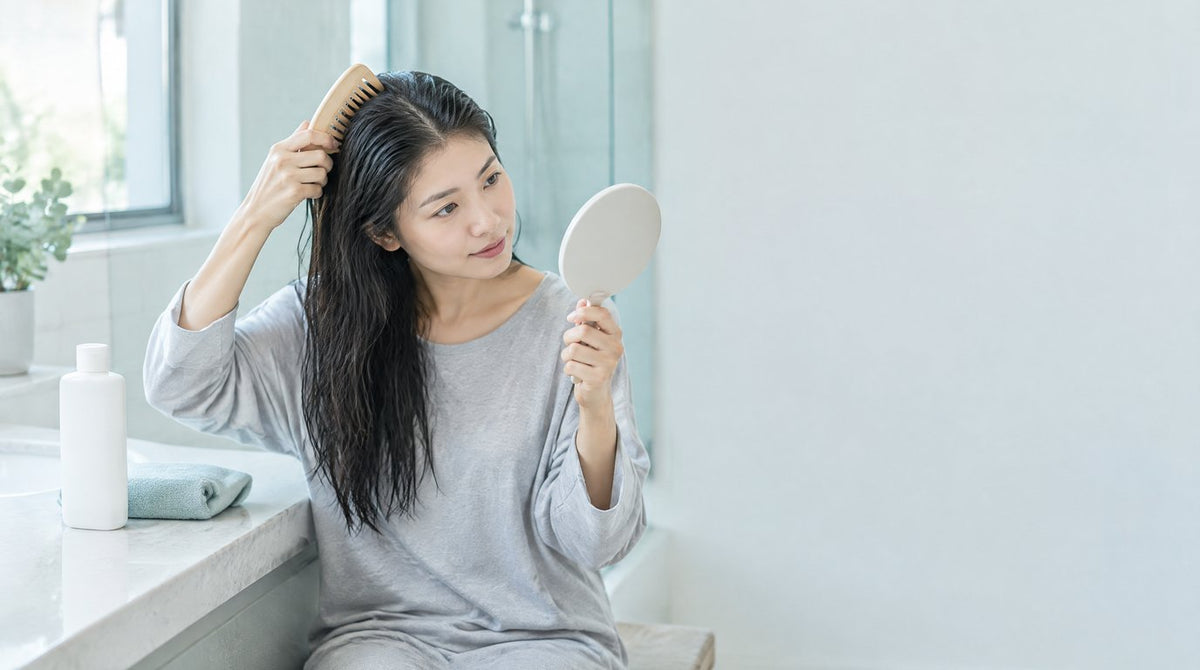 A woman combing her damp hair and checking her scalp in a hand mirror