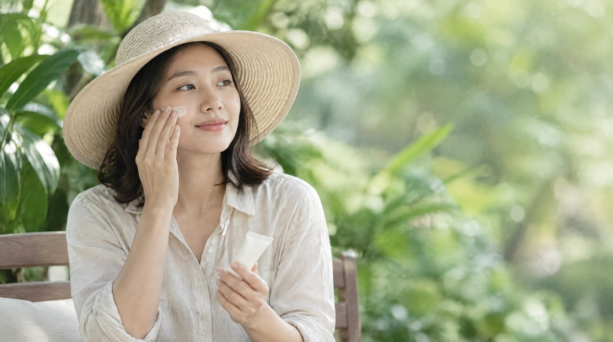 A woman in a sun hat applying sunscreen to her face in a shaded garden