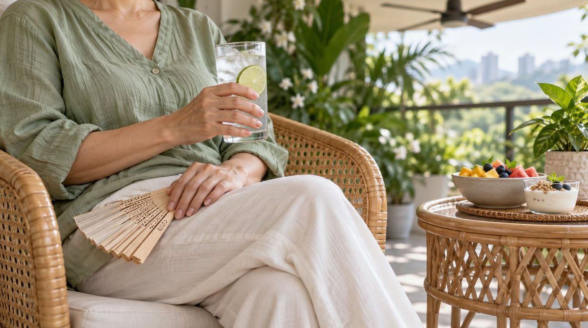 Midlife woman in a sage green blouse relaxing on a shaded balcony with a glass of iced water and a hand fan, beside fruit and yoghurt