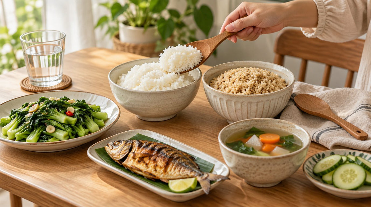 Bowls of white rice and brown rice with stir-fried sawi, grilled fish, vegetable soup and cucumber on a wooden table