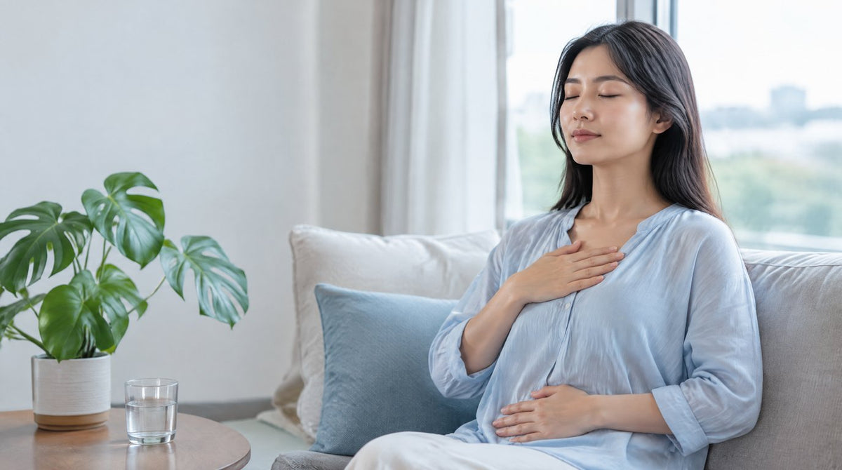 A woman sitting on a sofa by the window with her eyes closed, practising slow breathing with one hand on her chest
