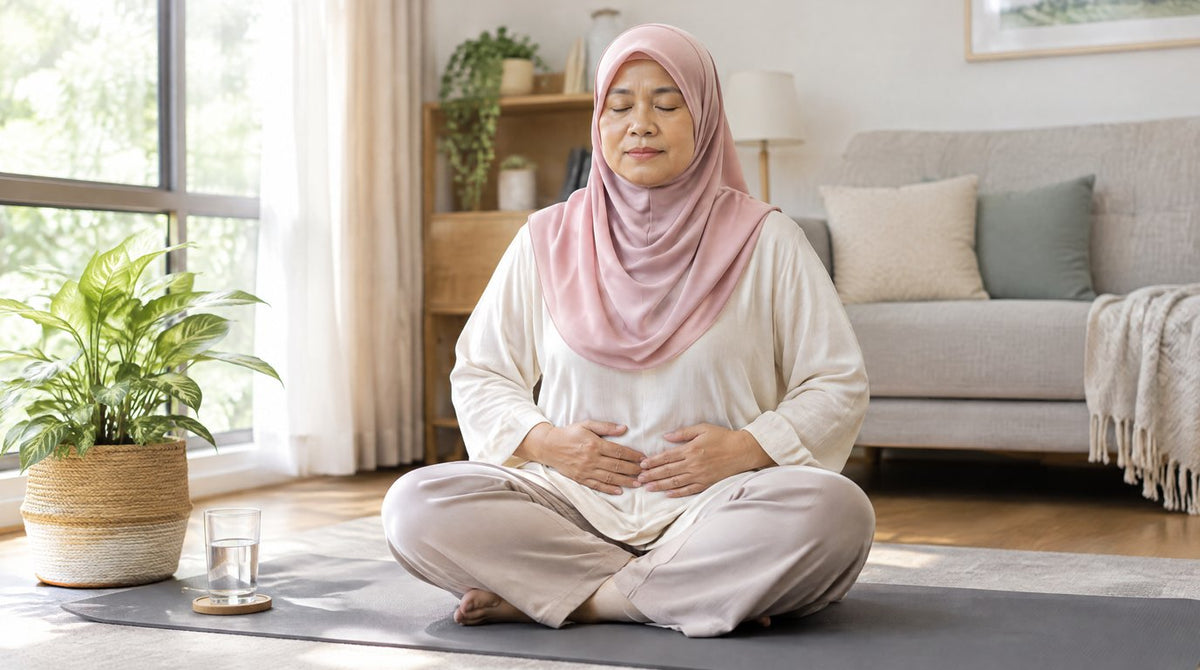 A Malaysian woman in a pink hijab sitting on a yoga mat at home, doing a calm breathing exercise with her hands on her lower abdomen