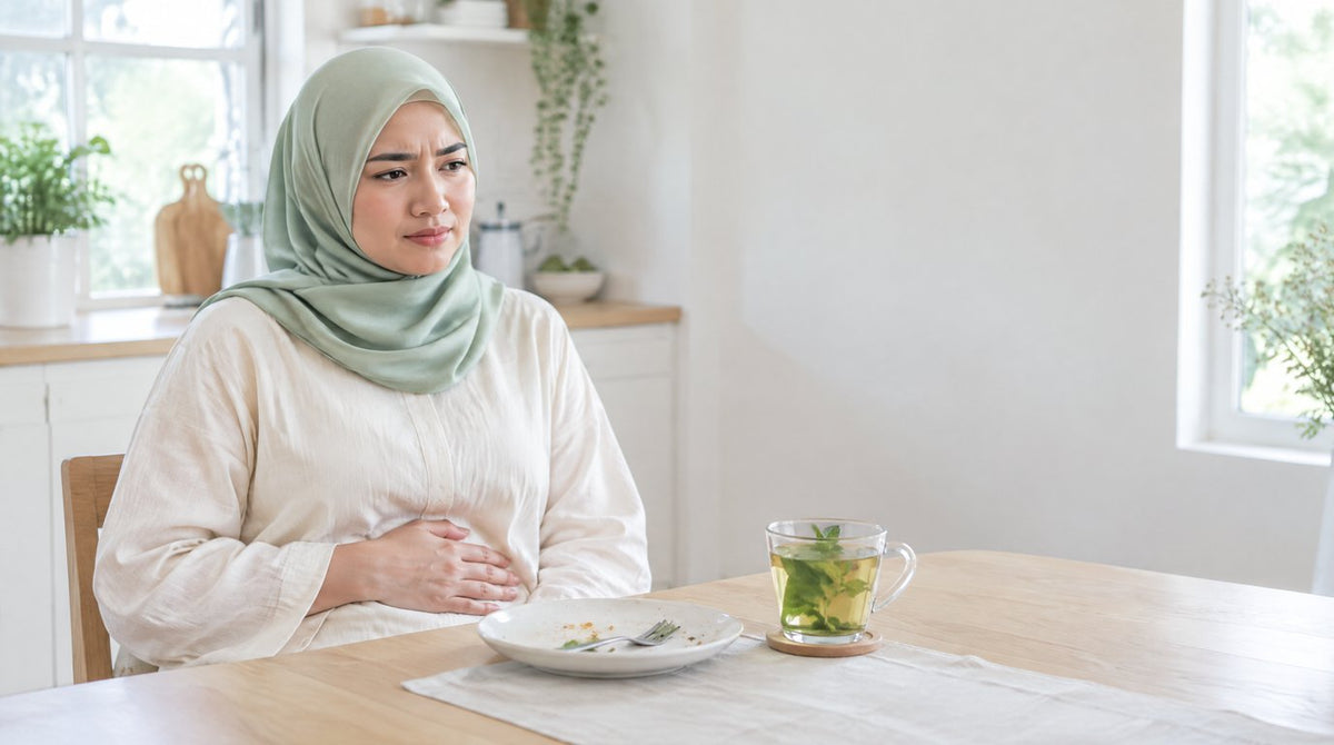 A woman in a hijab at a dining table with her hand on her tummy after a meal, with a cup of peppermint tea
