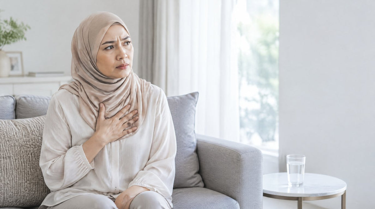 A woman in a hijab sitting on a sofa with her hand on her chest, a glass of water on the table beside her