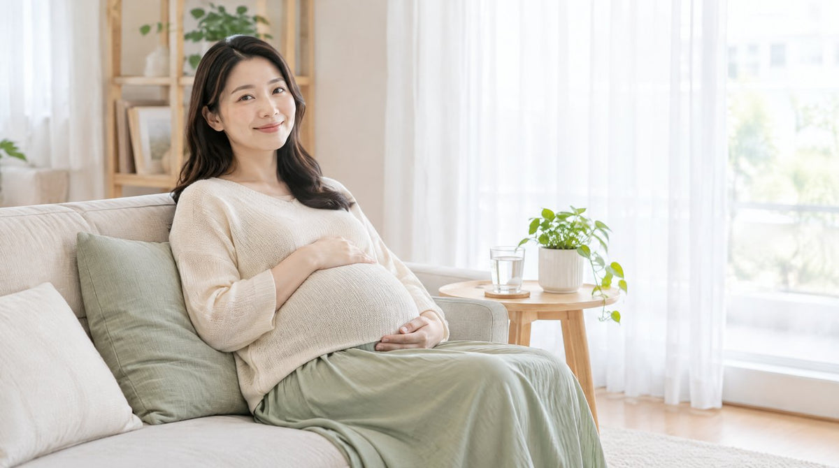 A smiling pregnant woman in a loose knit top and long skirt resting her hand on her bump while sitting on a sofa at home
