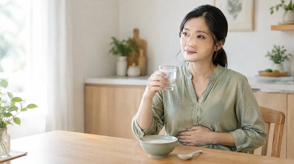 A woman at a kitchen table holding a glass of warm water with a hand resting on her tummy, beside a bowl of porridge